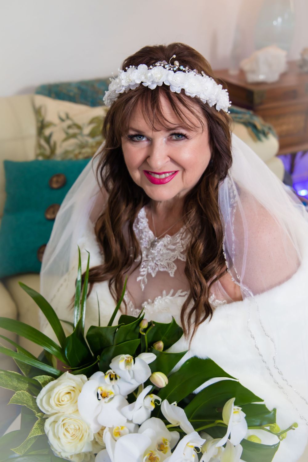 A bride in a white wedding dress and veil poses with a bouquet of white roses and greenery in a casual indoor setting.