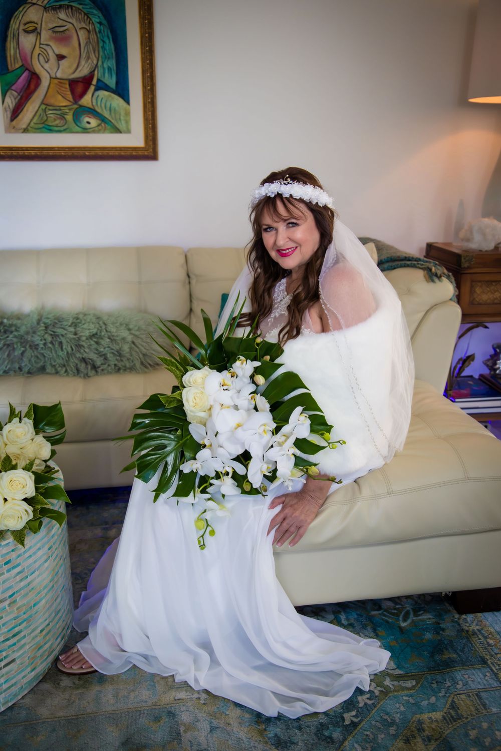 A person in a white wedding dress sits on a beige couch with a lush white floral bouquet.