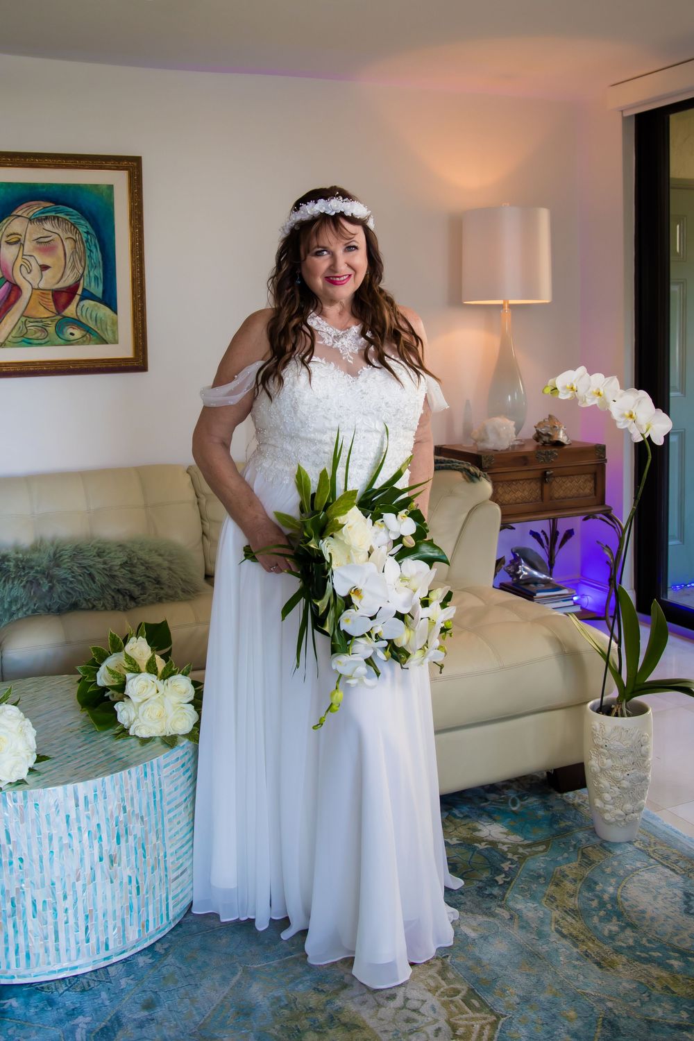 A person in a flowing white dress holds a bouquet of white flowers while standing in an elegant room with blue carpet.