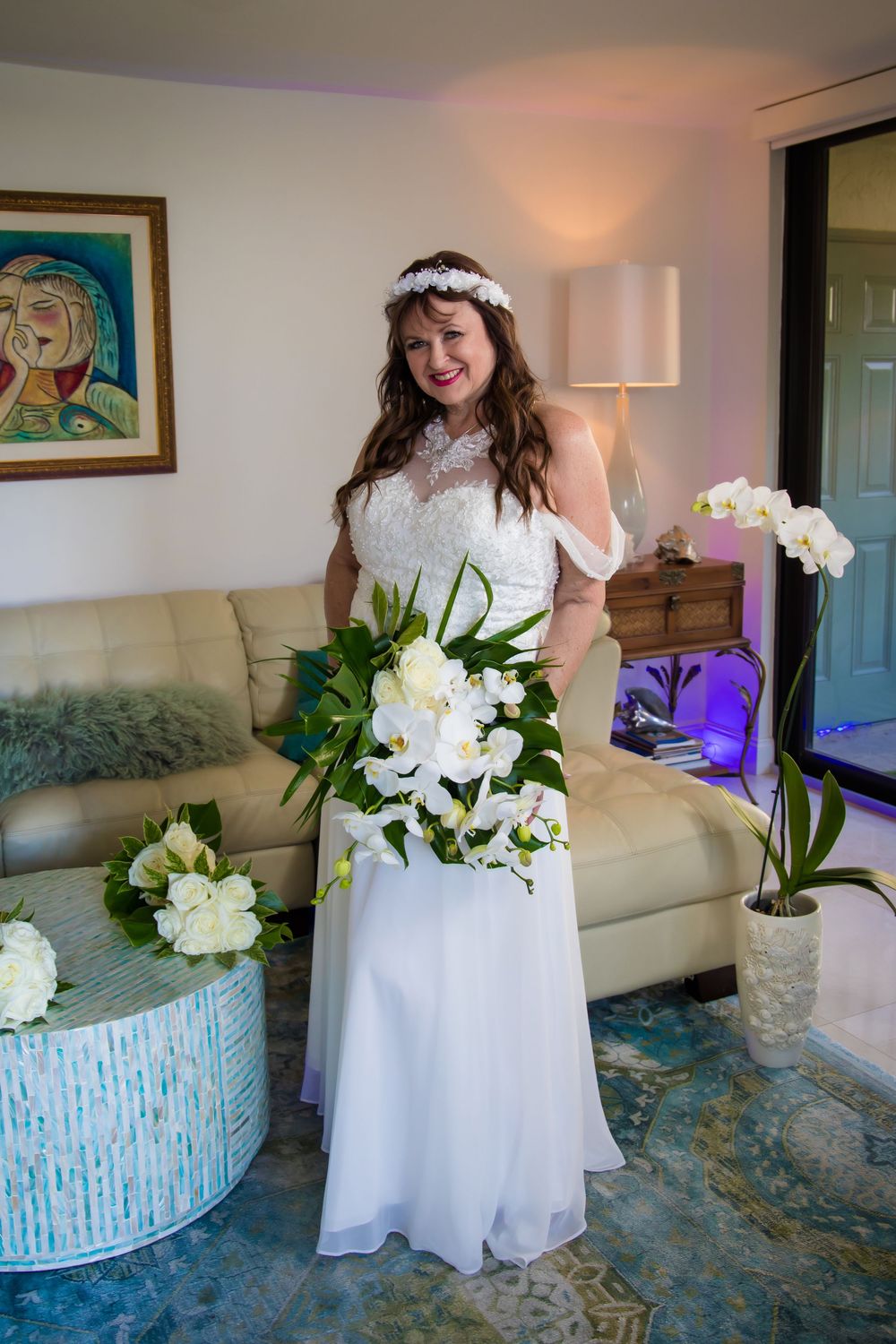 A bridal figure in a white lace dress stands beside elegant white floral arrangements in a softly lit room.
