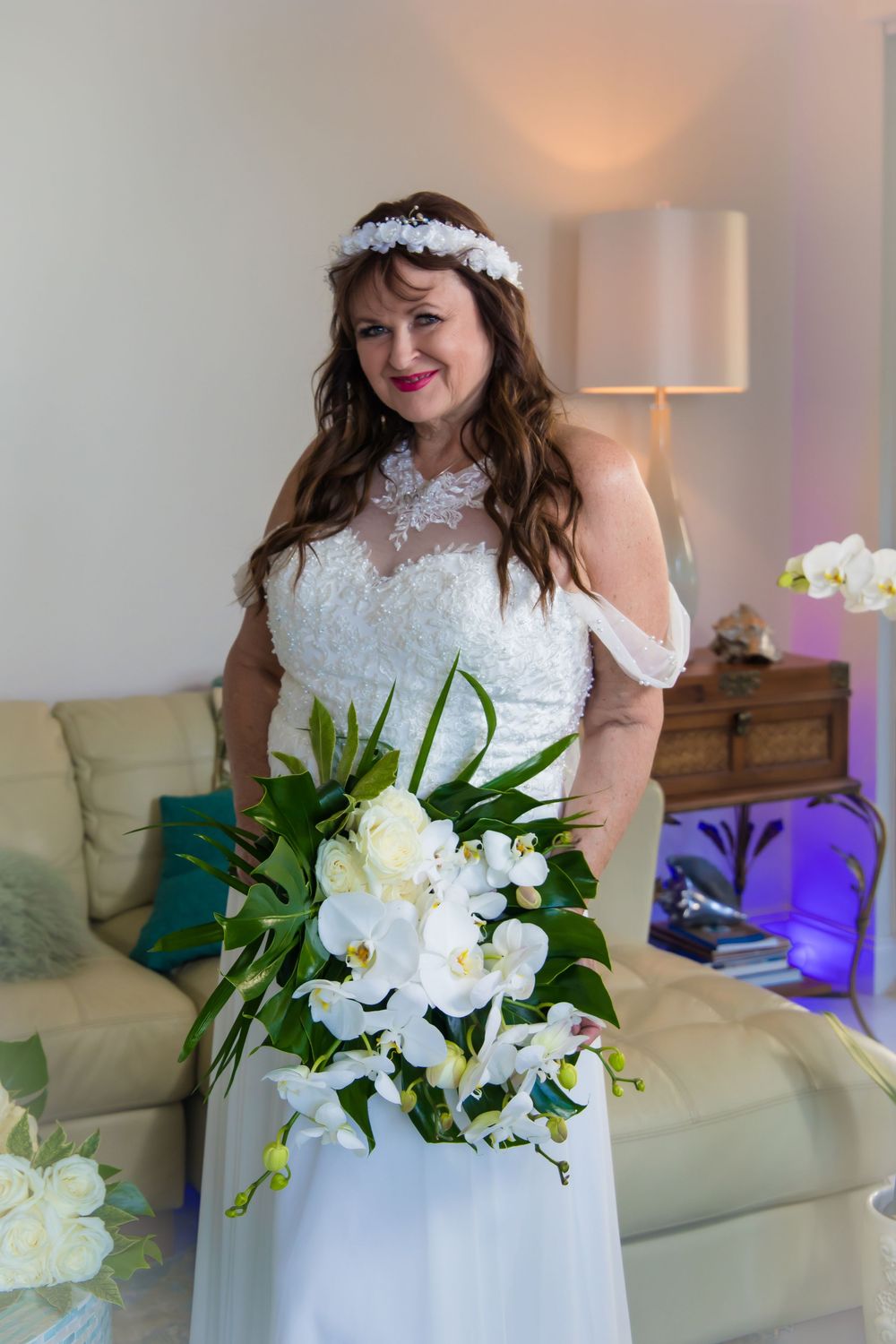 A bride in a white lace wedding dress holds a beautiful bouquet of white lilies and greenery in an indoor setting.