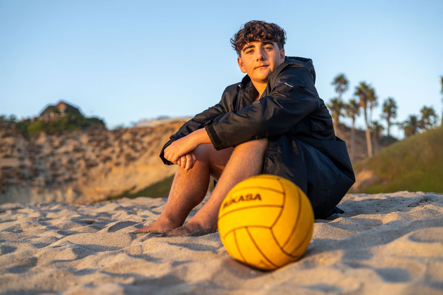 A person sits on sandy beach holding a yellow volleyball during golden hour lighting.