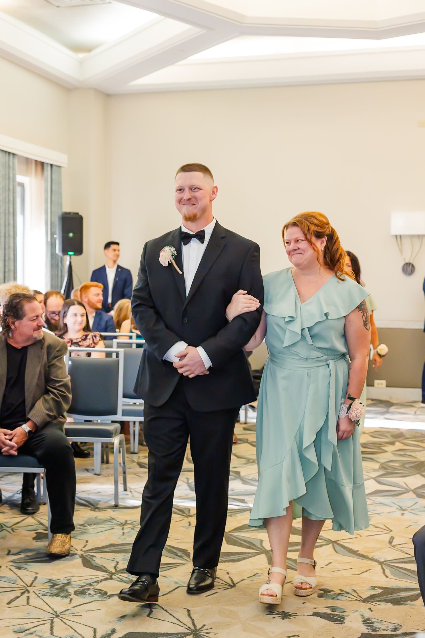 Wedding party members in formal attire dance together in a hotel ballroom with patterned carpet.