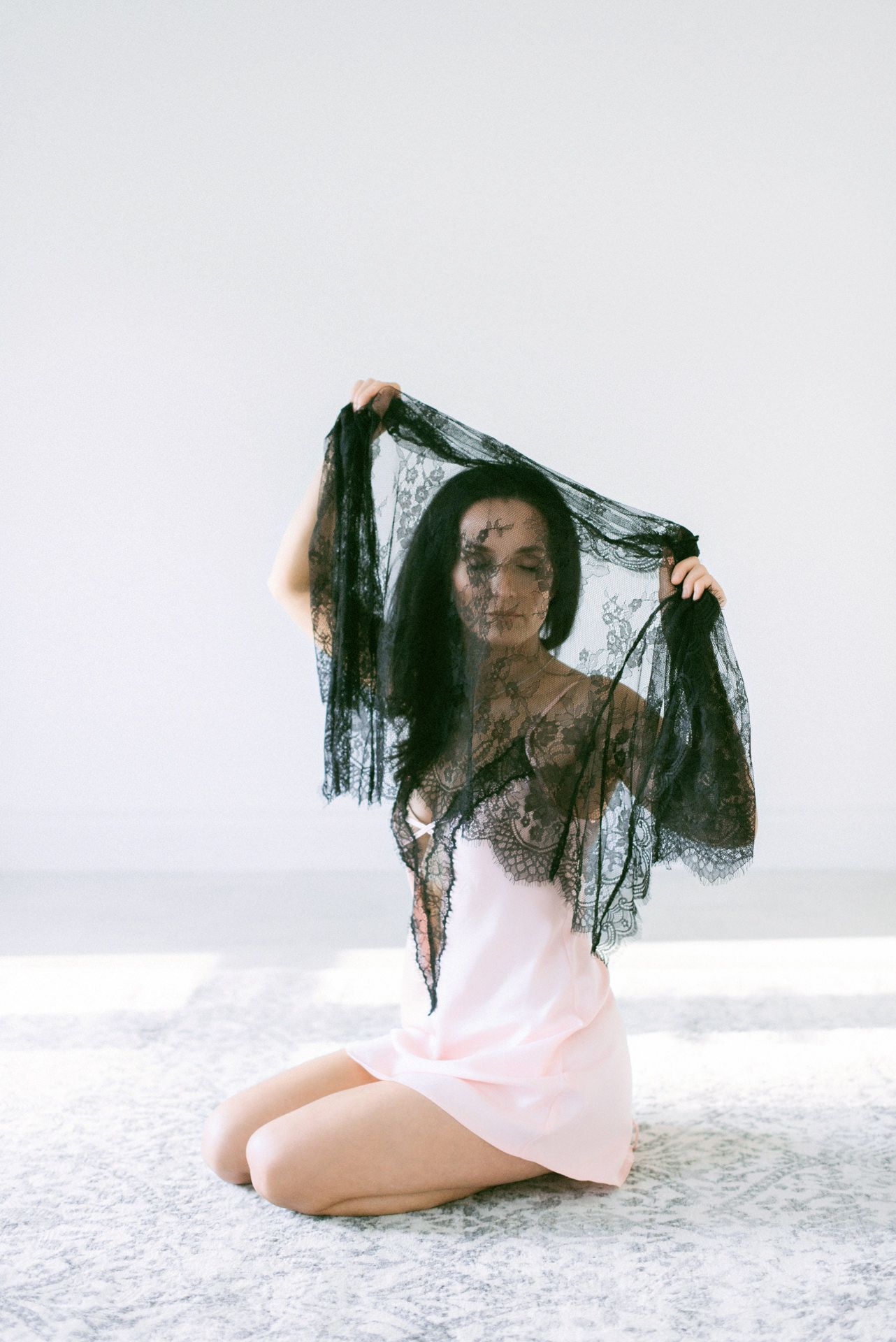 Figure in pink dress kneeling while holding sheer black lace veil against white background in artistic portrait.