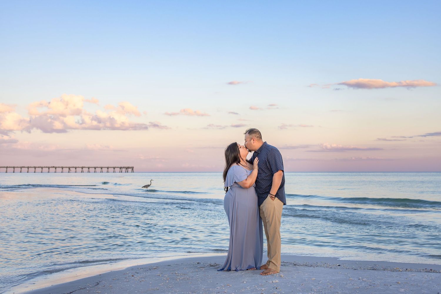 A couple stands together on a beach at sunset with a wooden pier in the background and gentle waves rolling in.