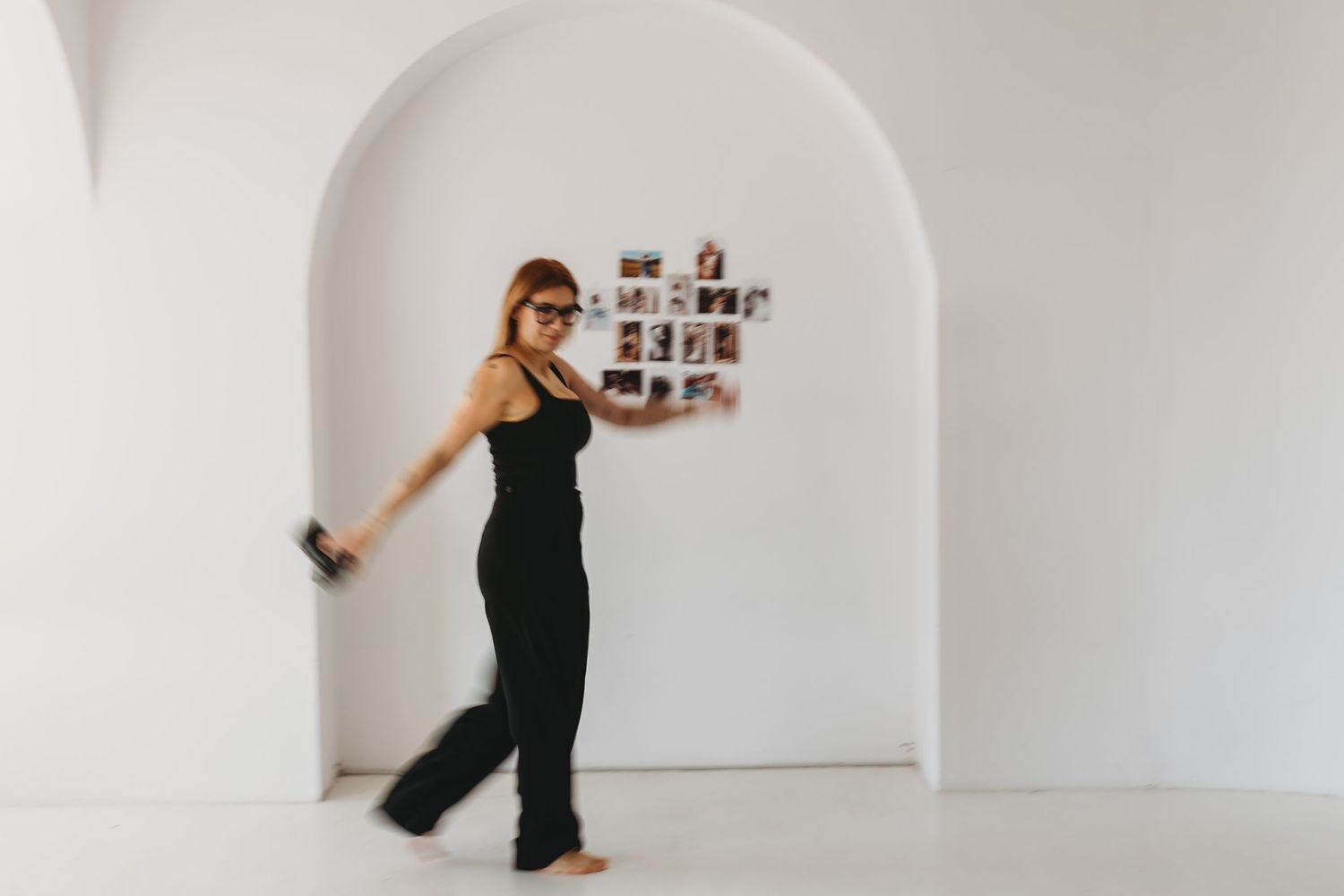 Someone in a black jumpsuit dances in a modern white room with an arched alcove and small photo gallery display.
