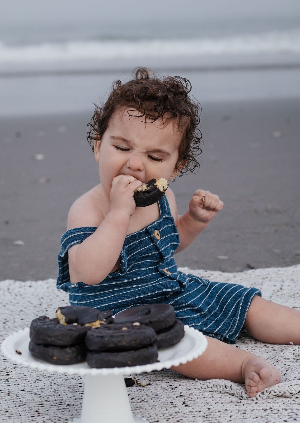 Child in striped outfit enjoys chocolate cookies while lying on beach blanket.