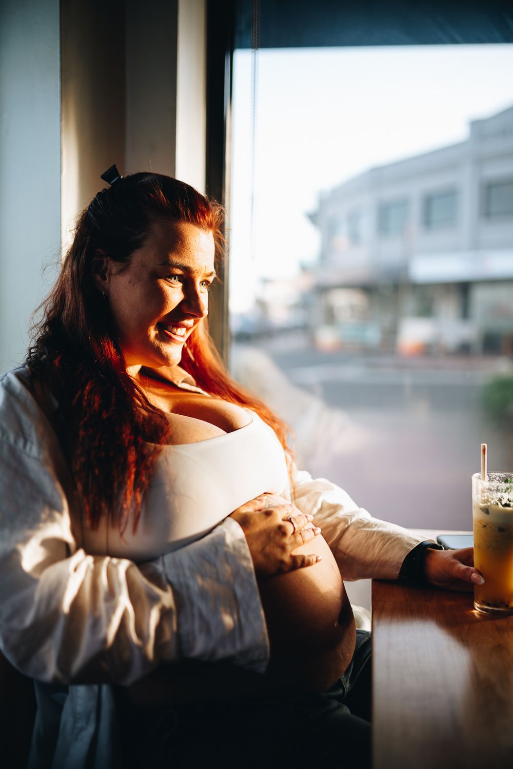 A smiling pregnant lady touching her belly in a cafe with sunlight through the big window taken by a Melbourne maternity photographer.