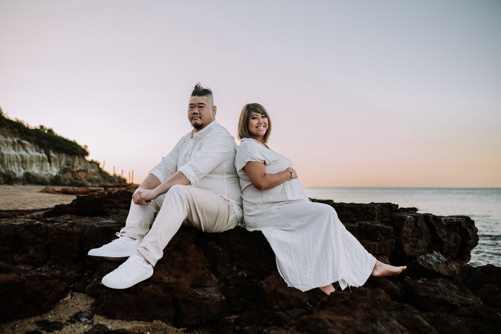 Pregnant couple sitting on a rocky formation on the beach during a sunset beach maternity photoshoot in Melbourne