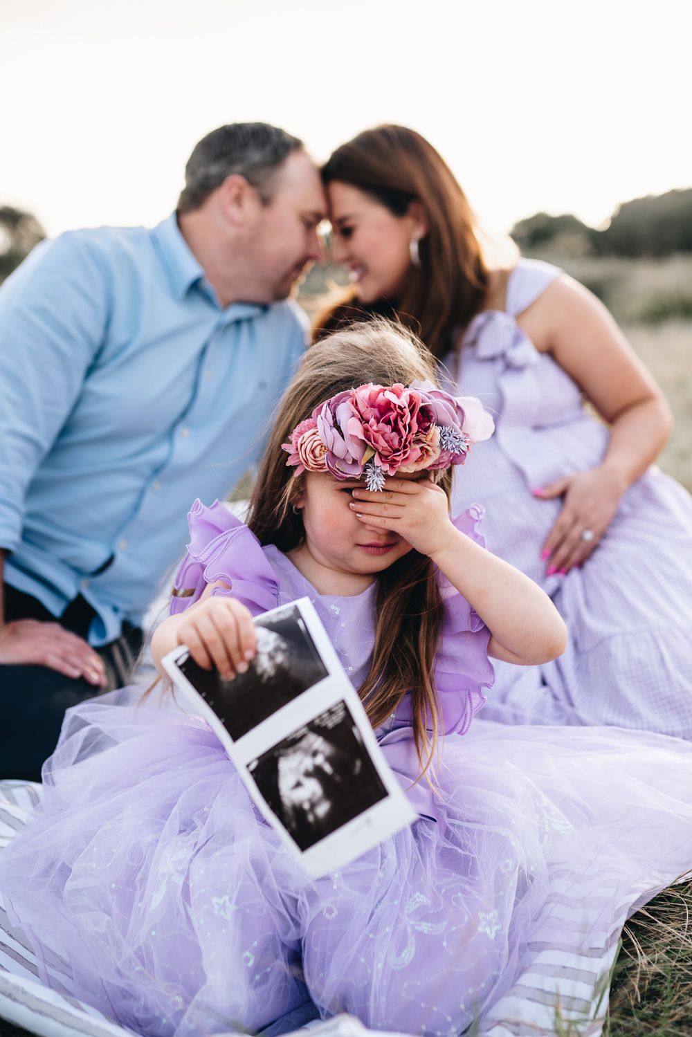 A toddler with an ultrasound picture of her unborn brother, while her parents are in the background by Melbourne maternity photographer
