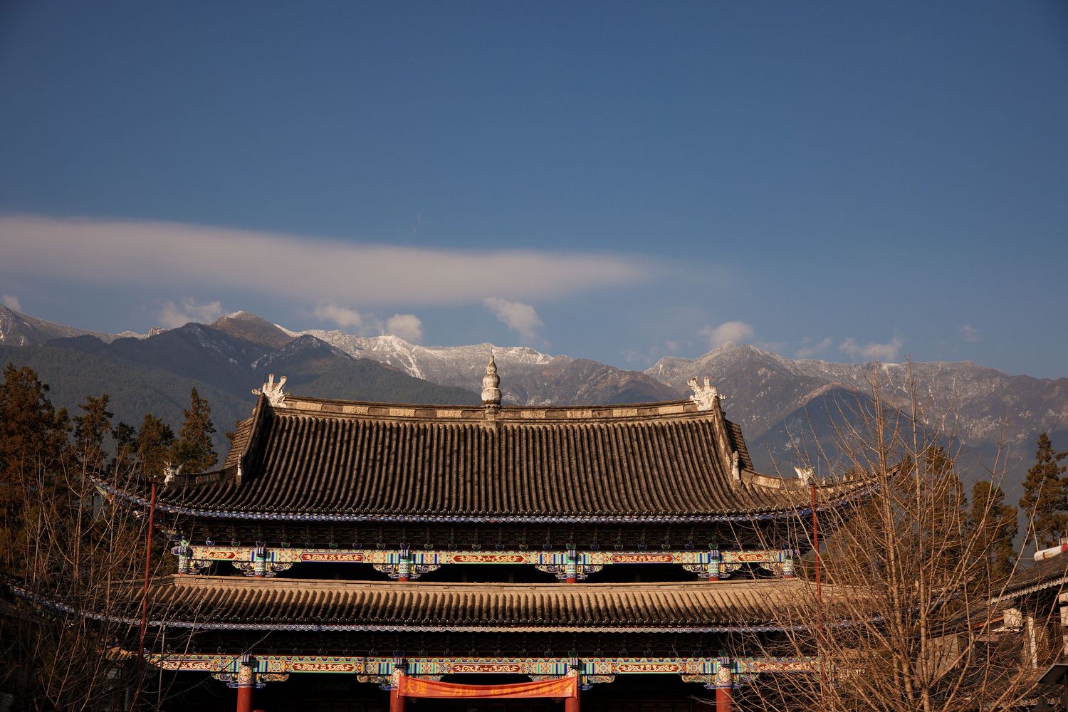 Traditional Chinese temple roof architecture set against snow-capped mountain peaks and blue sky in the background.