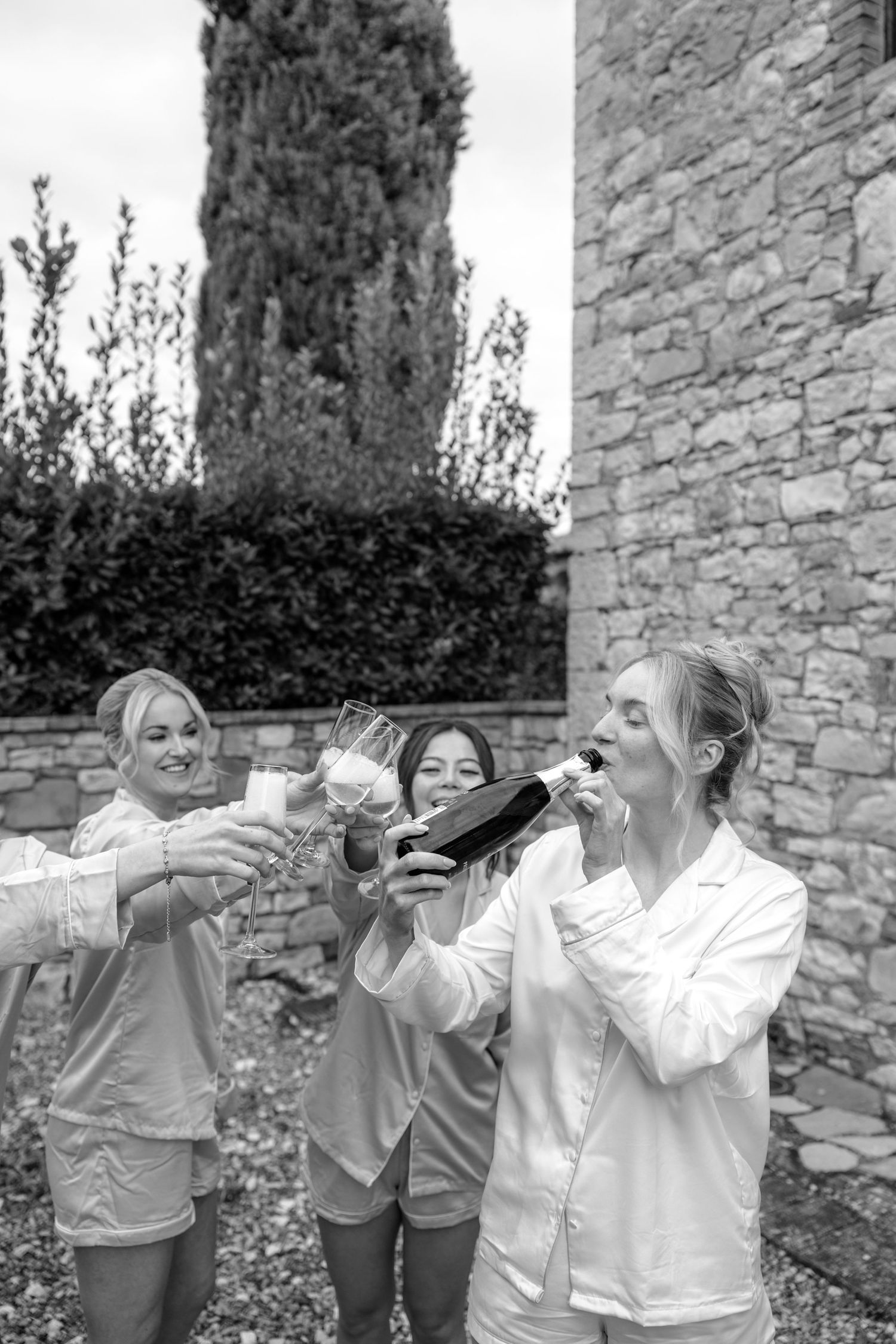 Bridesmaids in matching outfits toasting and drinking champagne against a stone wall.