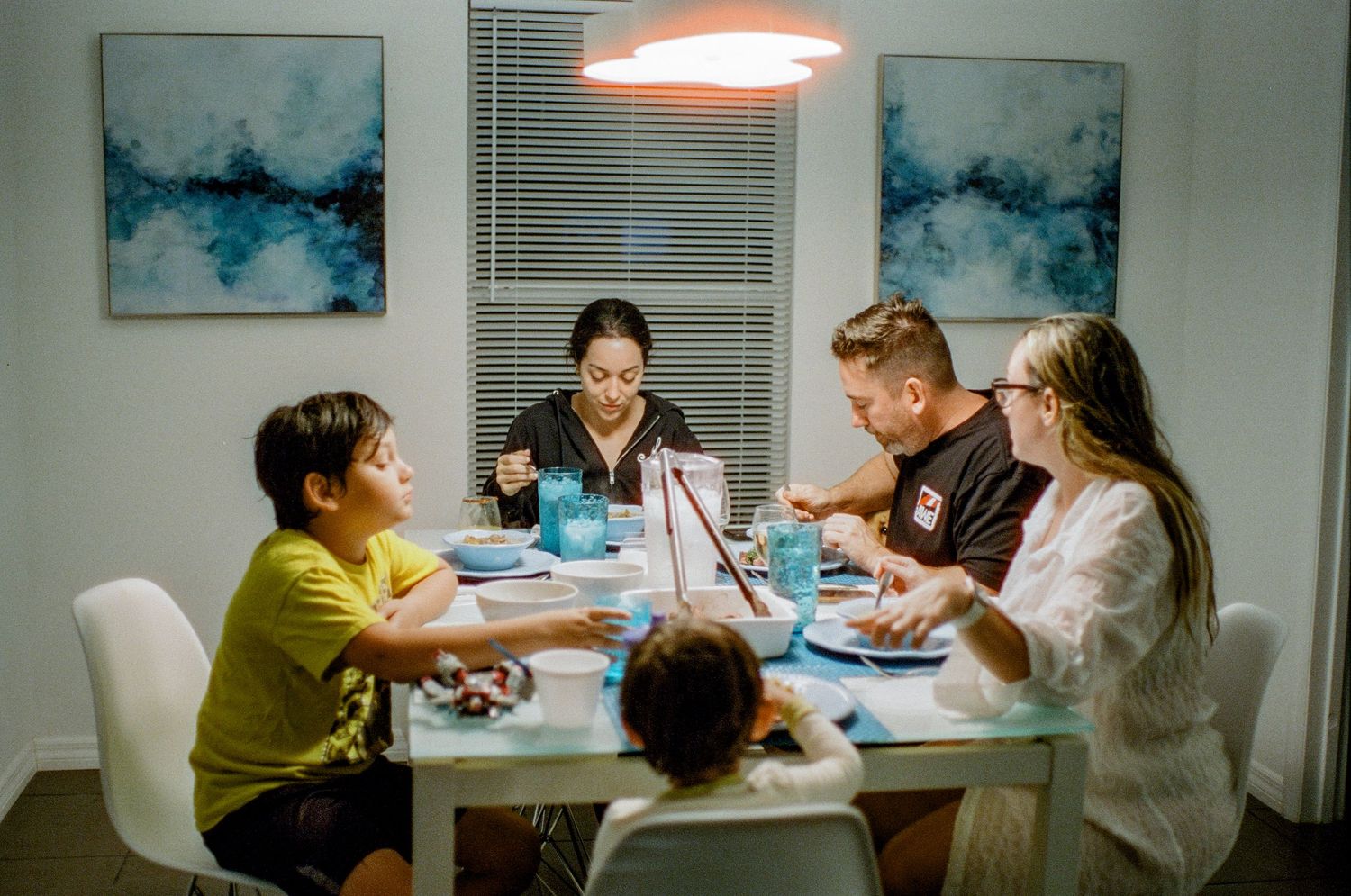 A group gathers around a dining table for a meal under warm lighting with blue abstract artwork on the walls.