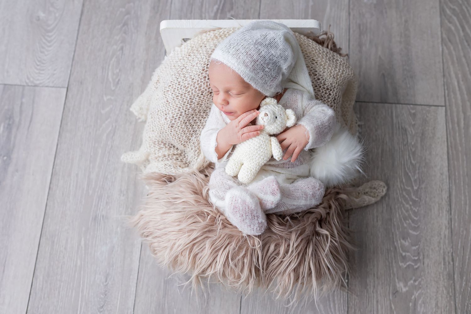Sleeping newborn in cozy white bonnet lies on beige fluffy blanket holding small plush toy on wooden floor.