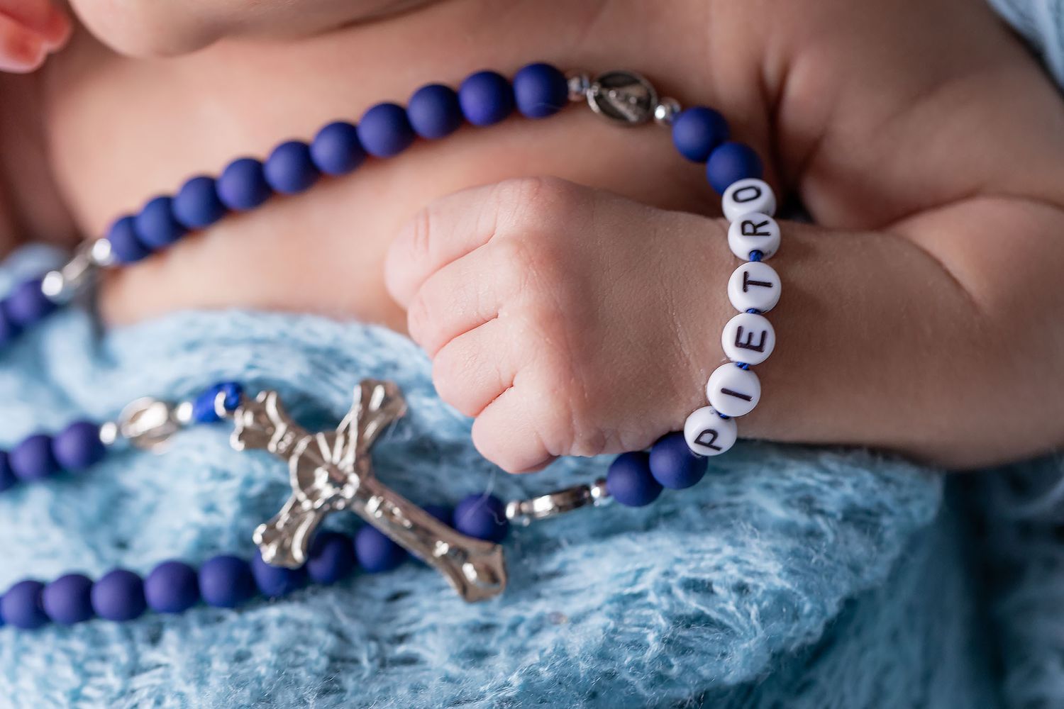 Baby's hand gently holds a blue and silver rosary bead necklace with cross pendant against soft blue fabric.