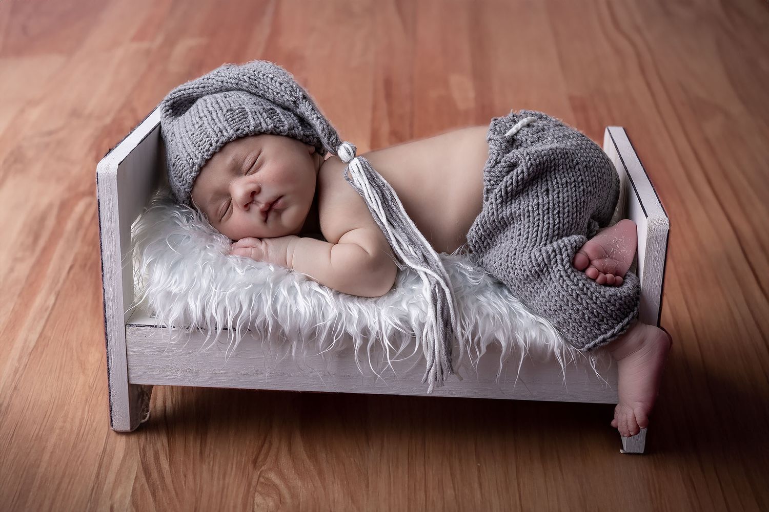 Sleeping newborn wearing gray knit outfit lies peacefully on white furry bedding in miniature wooden bed.