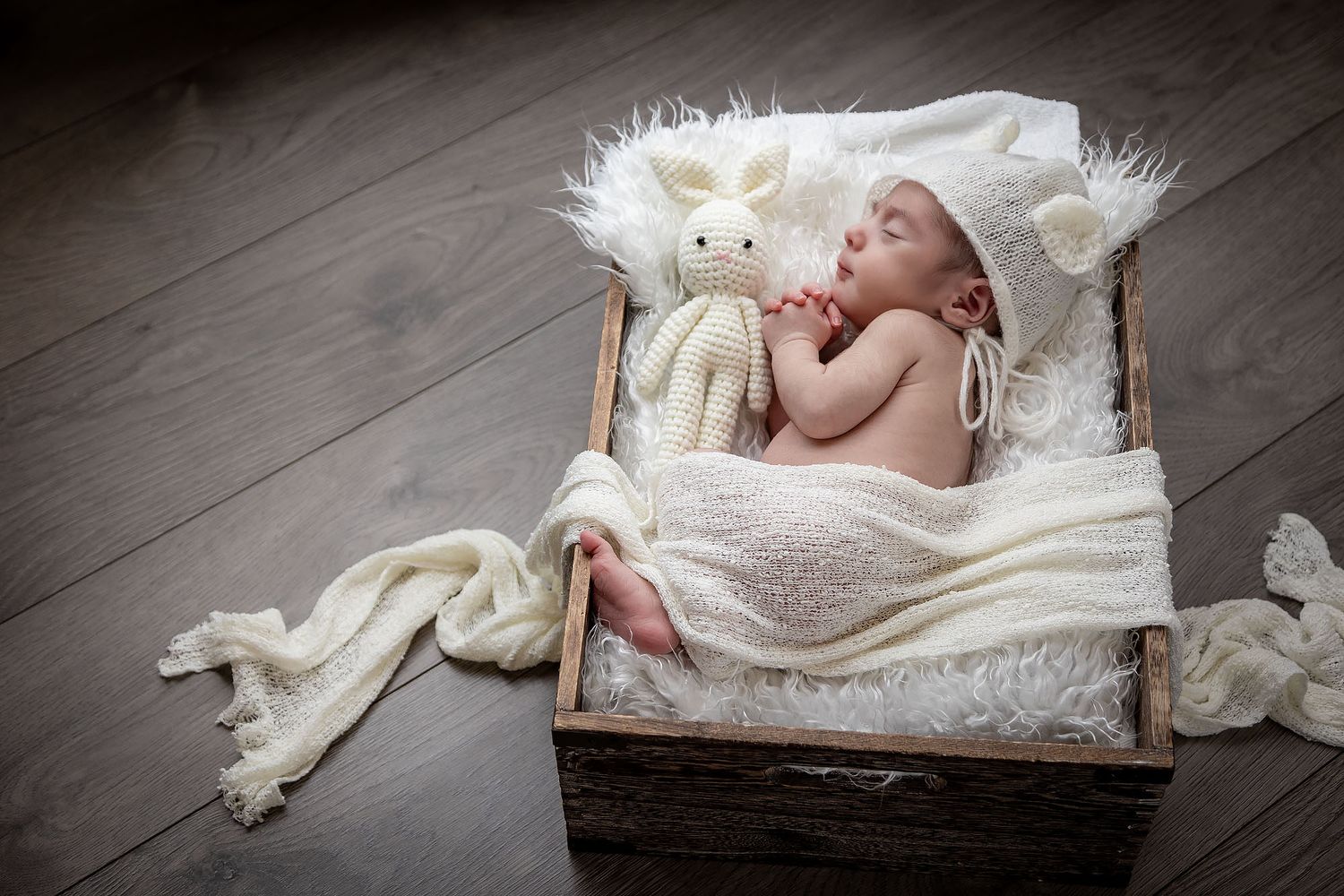 A sleeping newborn rests peacefully in a wooden box lined with soft white fur alongside a plush stuffed animal.