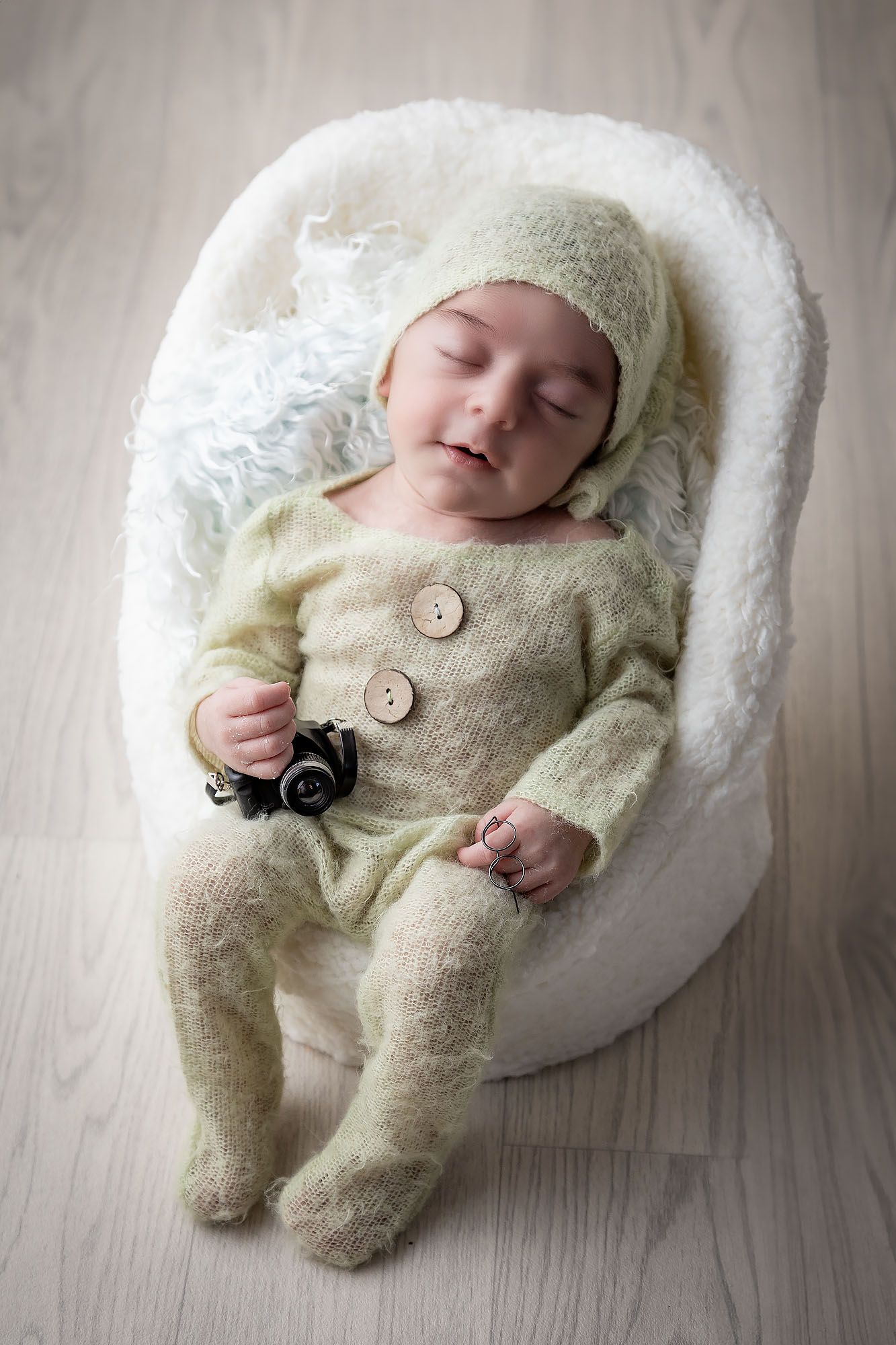 A sleeping infant wearing a cozy knit outfit and hat lies comfortably in a plush white chair on a wooden floor.