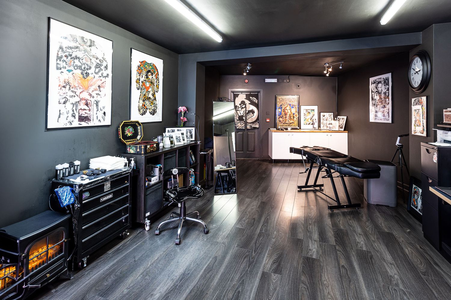 Modern tattoo studio interior with dark walls, hardwood floors, artwork displays, and professional equipment setup.