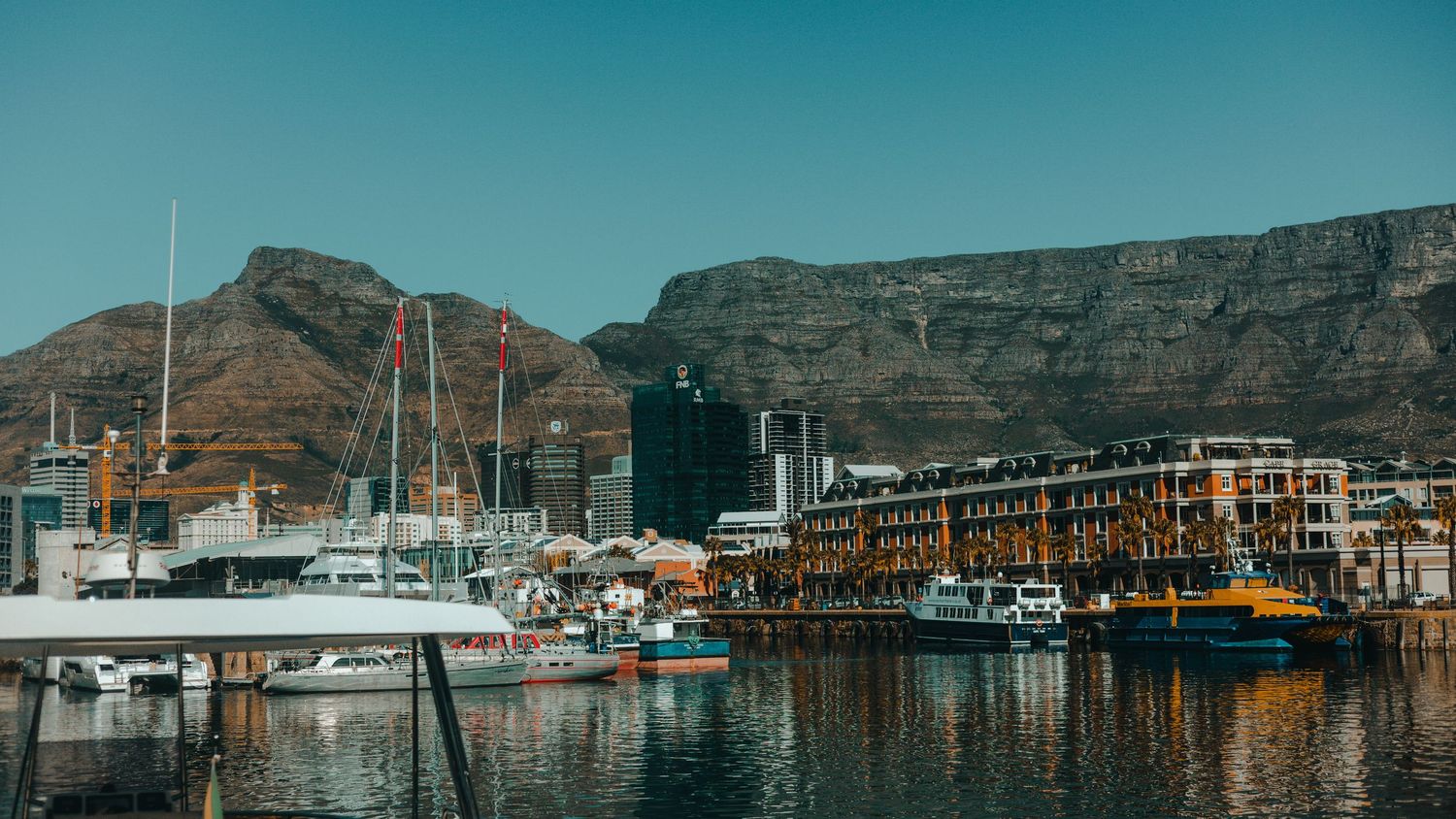 Wide panorama of Cape Town harbor with mountain backdrop and boats moored in blue water.