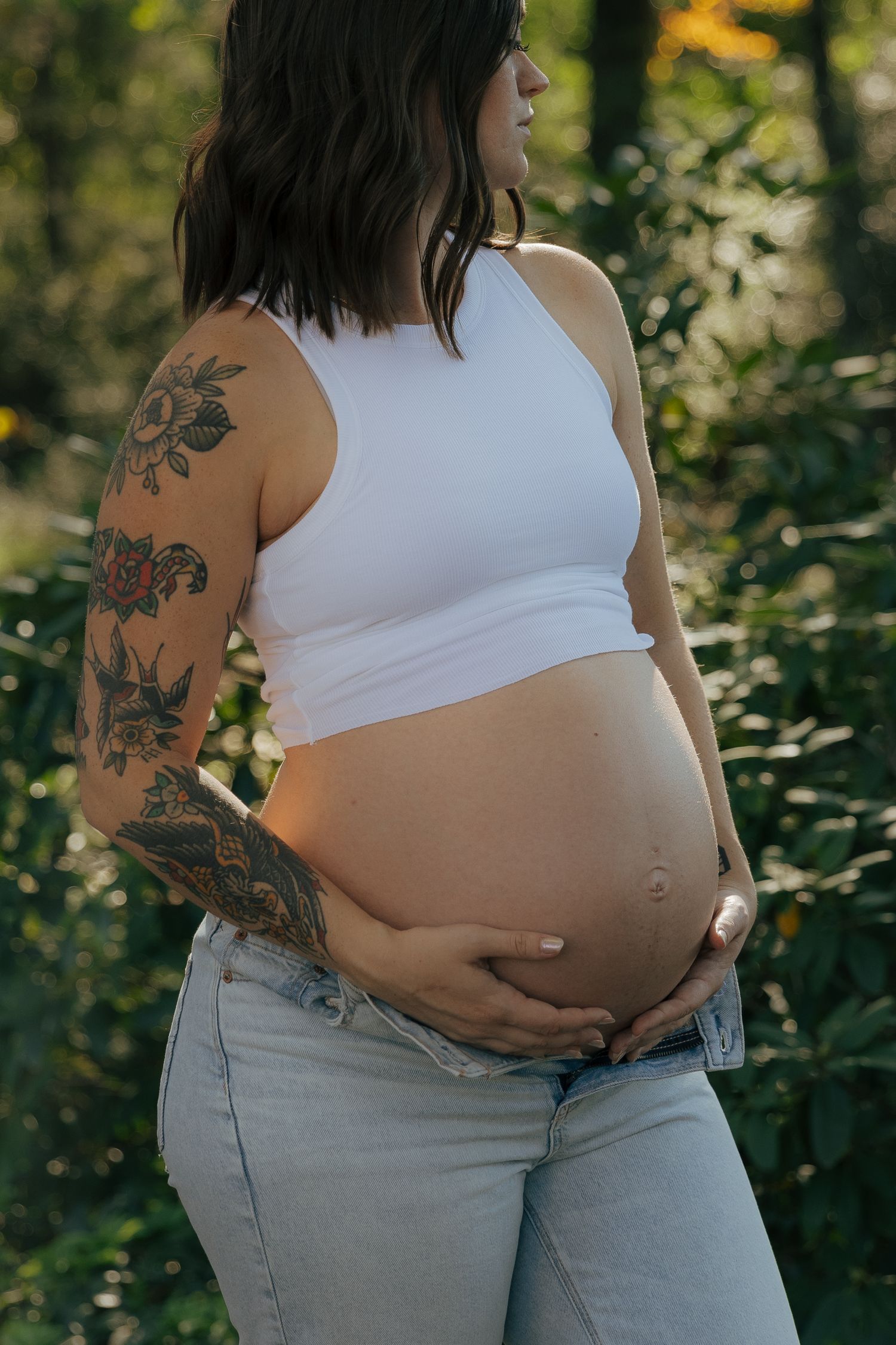 Pregnant woman in white tank top and jeans stands in sunlit forest while tenderly touching her neck.