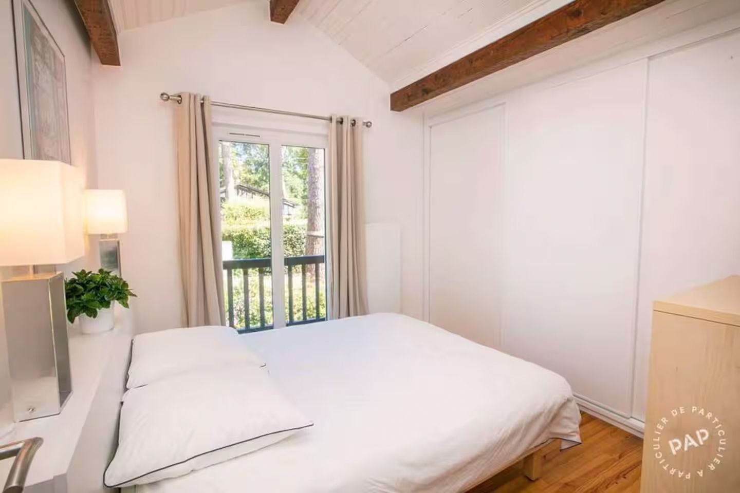 Cozy bedroom with white bedding, wooden ceiling beams, and glass doors leading to a private balcony with natural light.