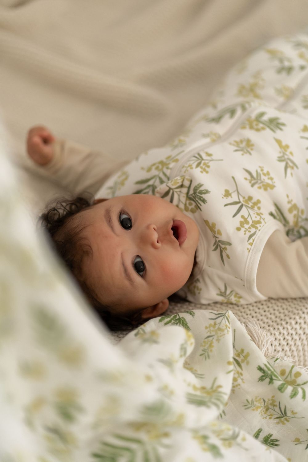 Close up of an alert baby lying on floral patterned bedding looking up at the camera.