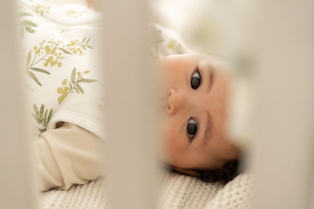 A small child rests peacefully looking up while laying on soft white bedding with delicate green leaves in view.