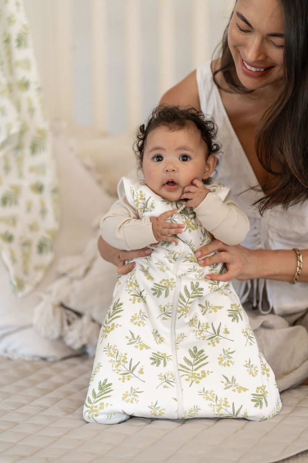 Baby wearing a floral sleep sack smiles while being held by caregiver on a bed with matching floral linens.