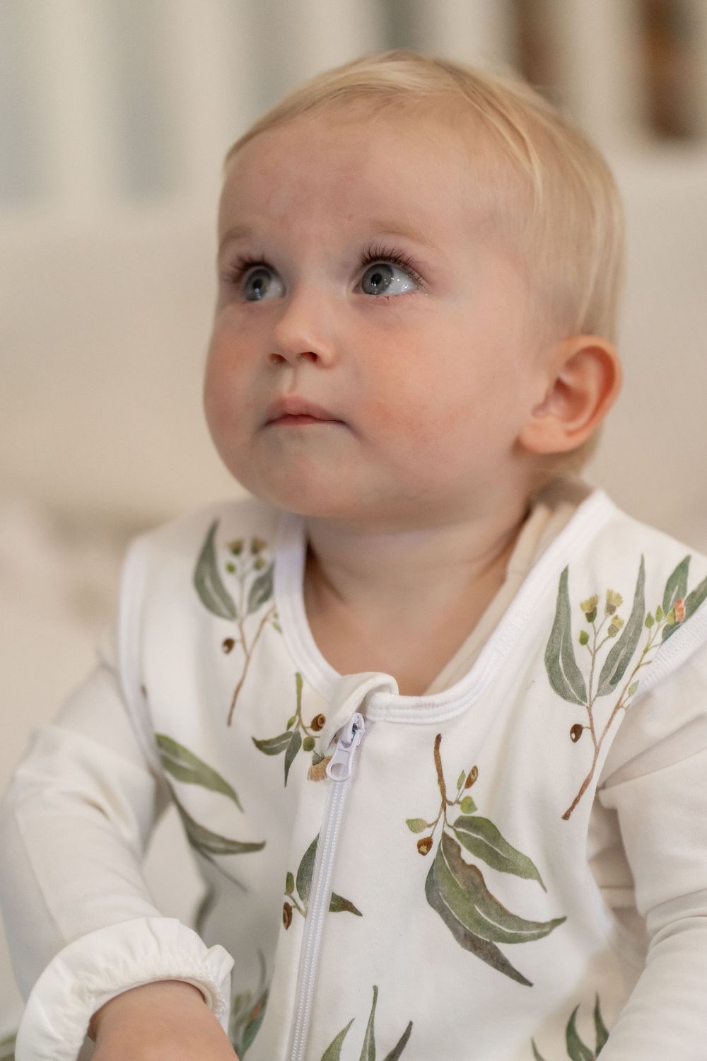 Sweet young child in a white zippered outfit with delicate green leaf and floral print pattern gazes upward pensively.