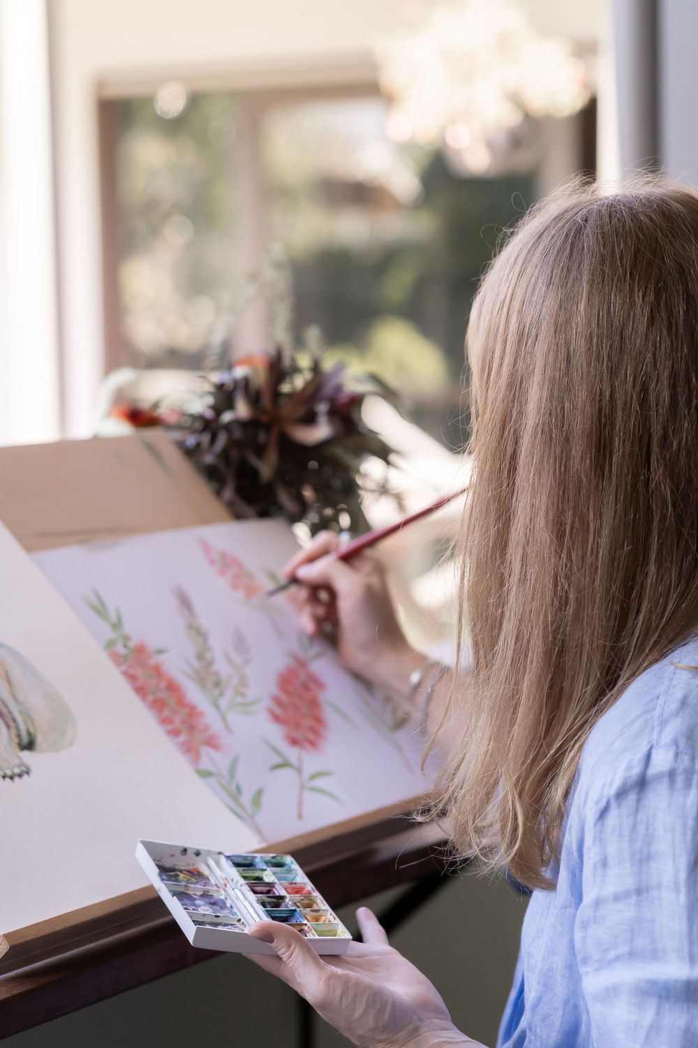 A person painting delicate floral watercolors at a desk near a window with natural light streaming in.