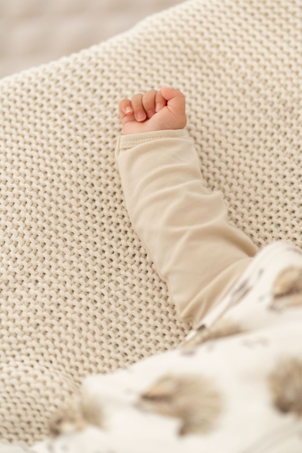 Small baby's fist and arm in cream colored sleeve resting on knitted blanket.