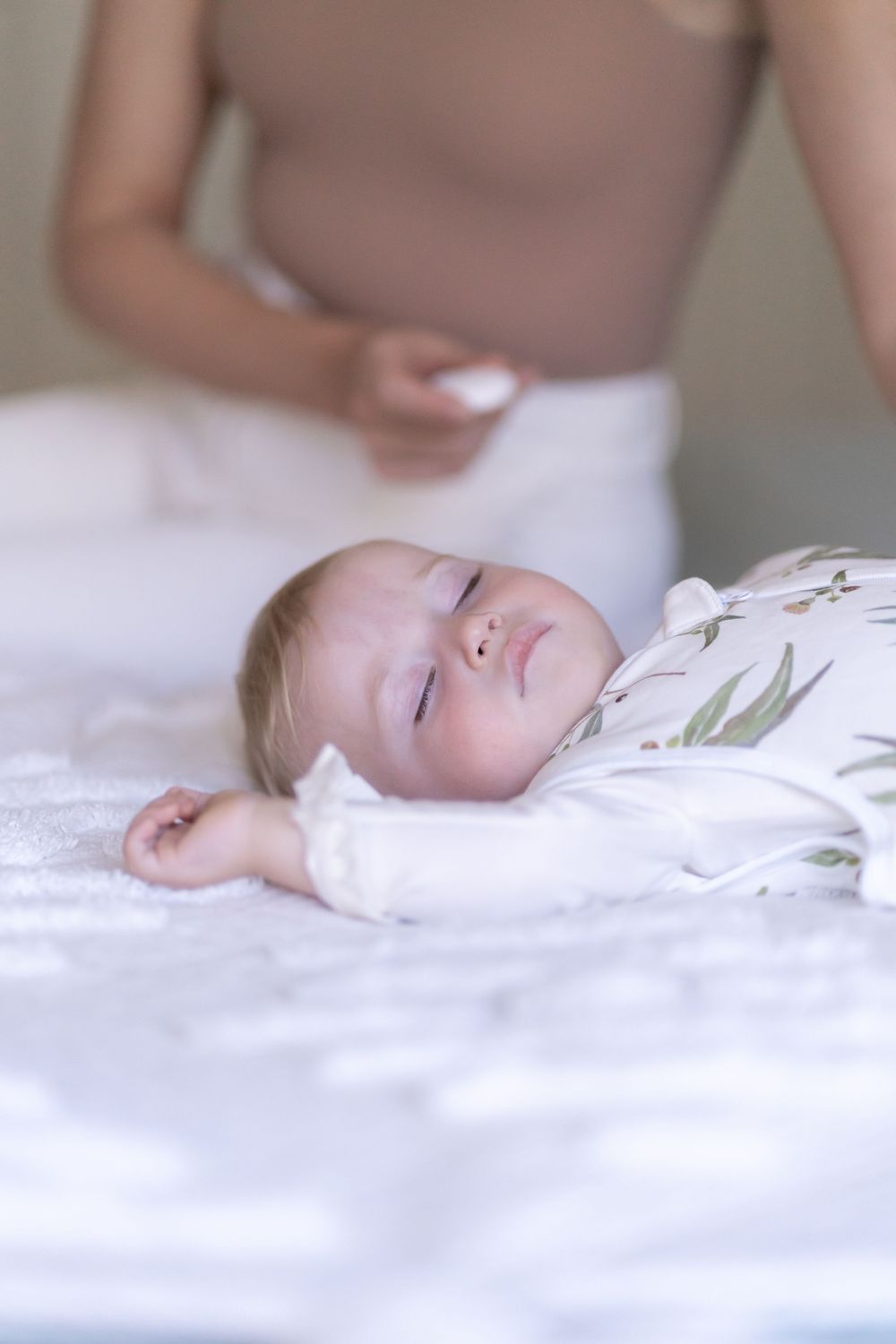 A peaceful baby sleeping on white bedding while wearing a floral patterned onesie.