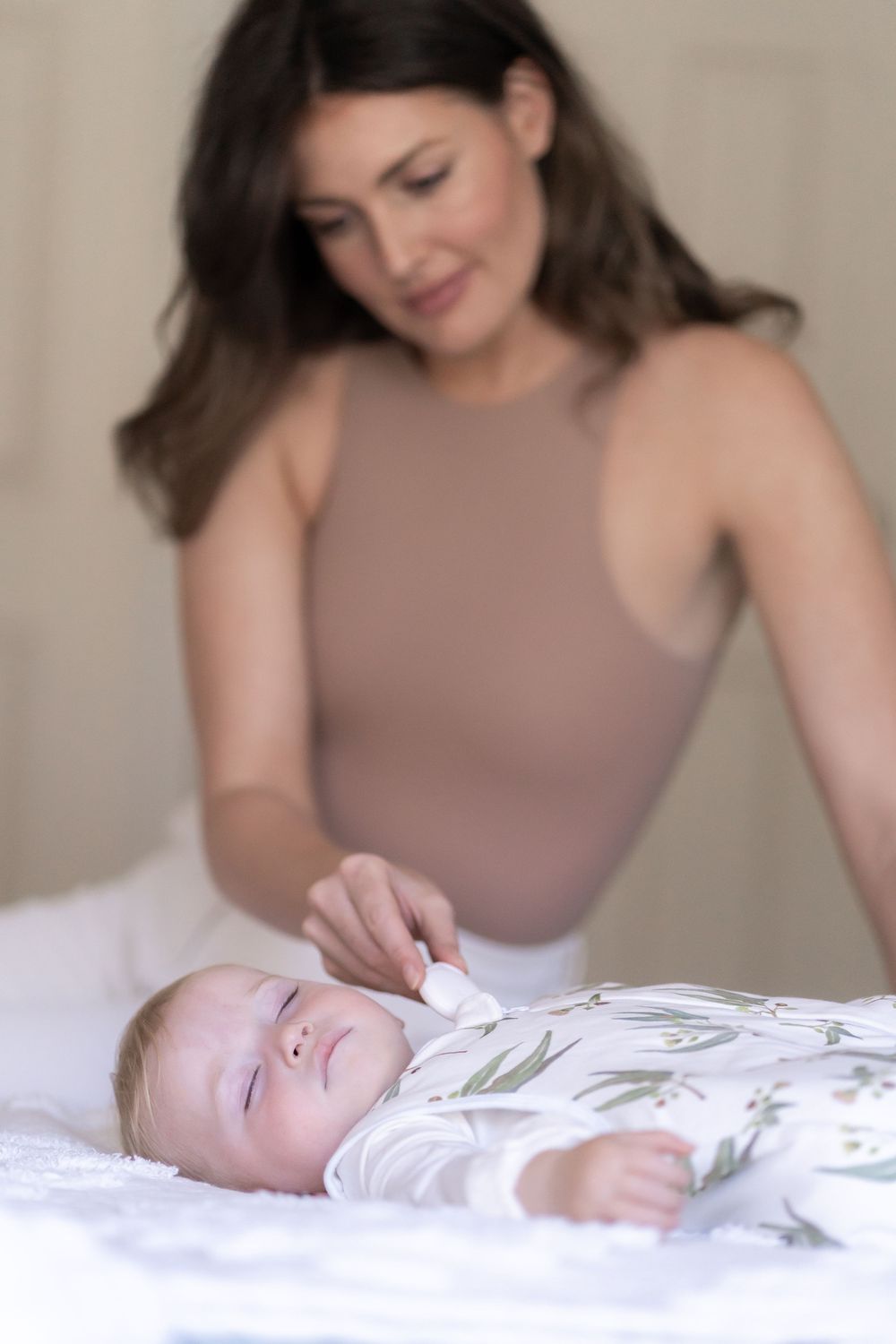 A caregiver gently touches a sleeping baby wrapped in floral bedding while lying on white linens.