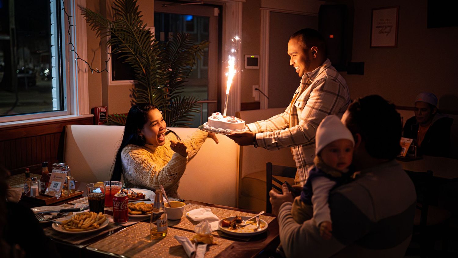 A family shares a warm candlelit dinner around a table in a cozy home setting with warm amber lighting.