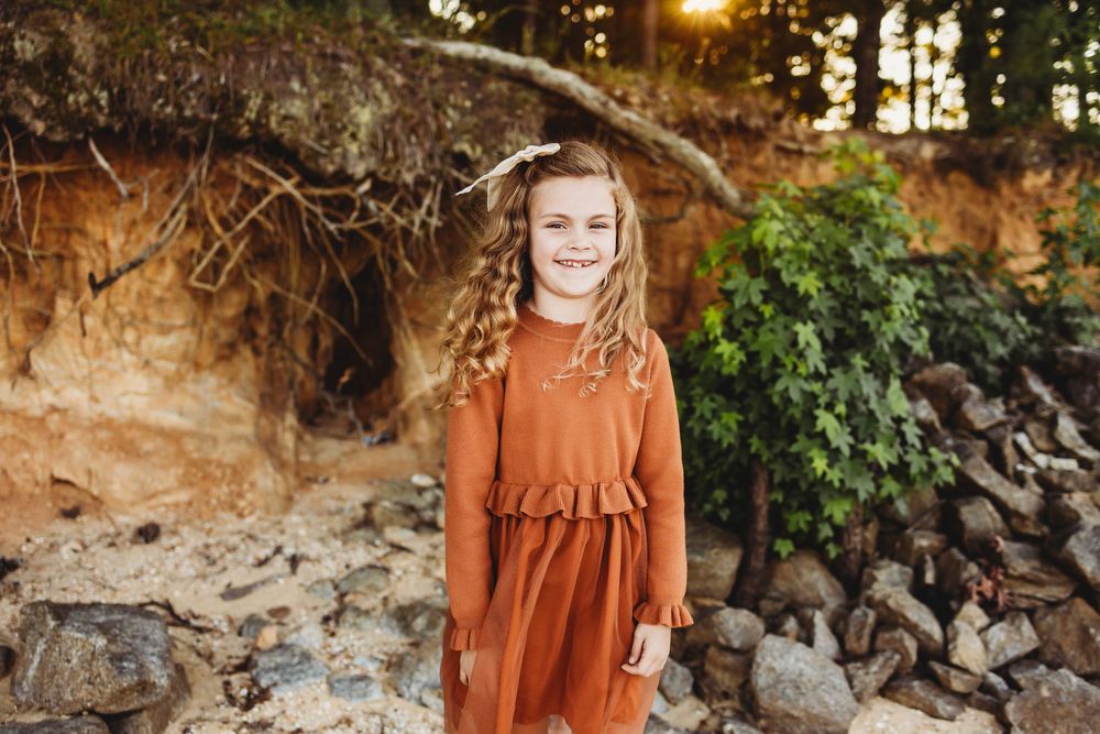 Child wearing a rust-colored dress stands in front of a natural rock wall and greenery during golden hour.