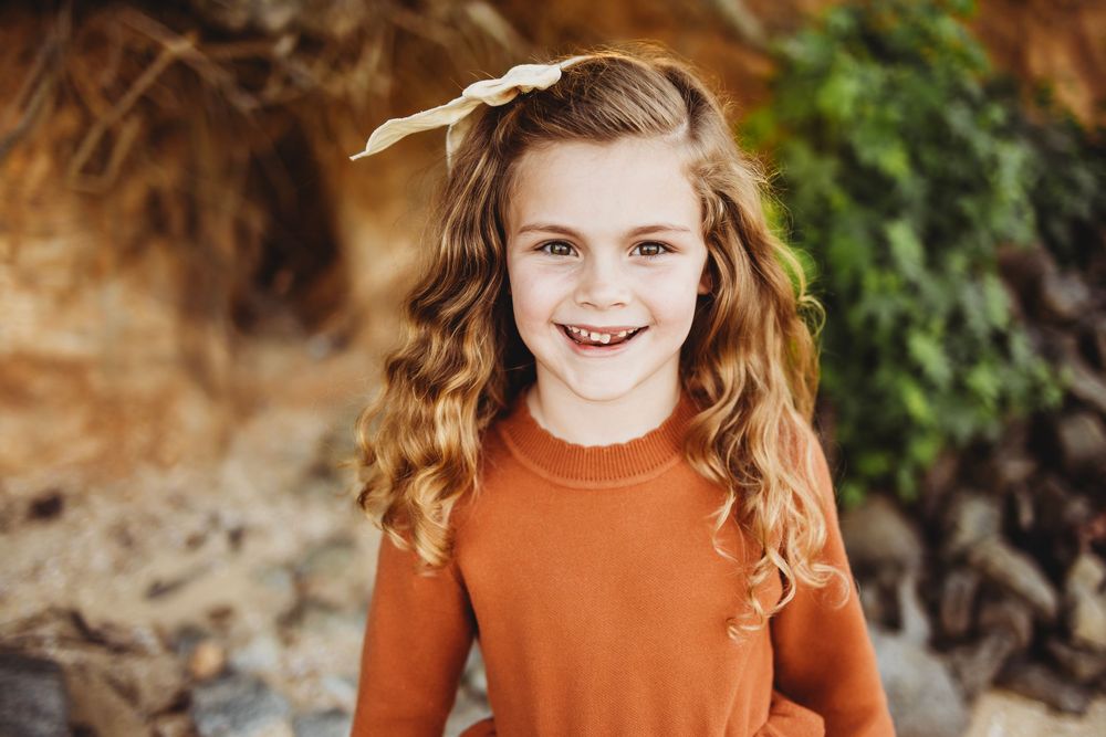 A young person in an orange sweater smiles warmly against an outdoor background with brown and green blurred foliage.
