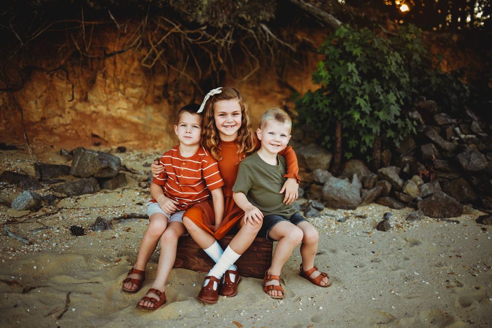 Three children in casual summer clothing sit together on sandy ground against a rocky outdoor backdrop.