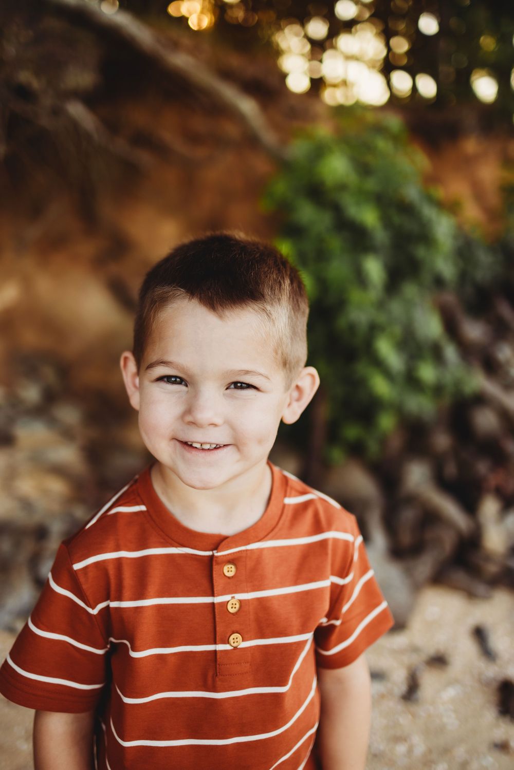 Smiling child wearing orange and white striped shirt outdoors with bokeh lighting and greenery in background.