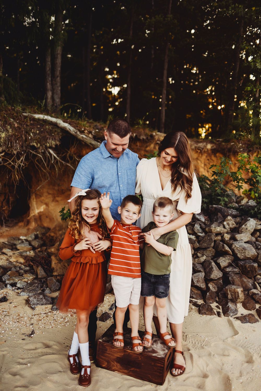 Group of five people standing together on a sandy beach area next to stone wall and trees at sunset.