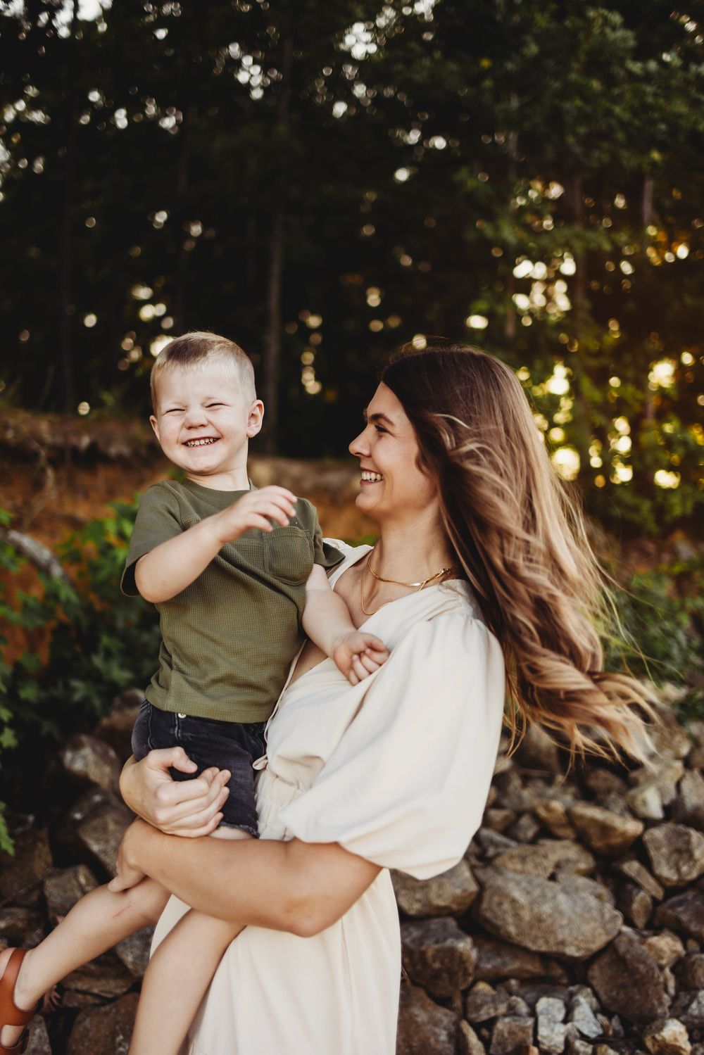 Joyful outdoor family portrait with white dress and olive shirt against natural stone wall and forest backdrop at sunset.