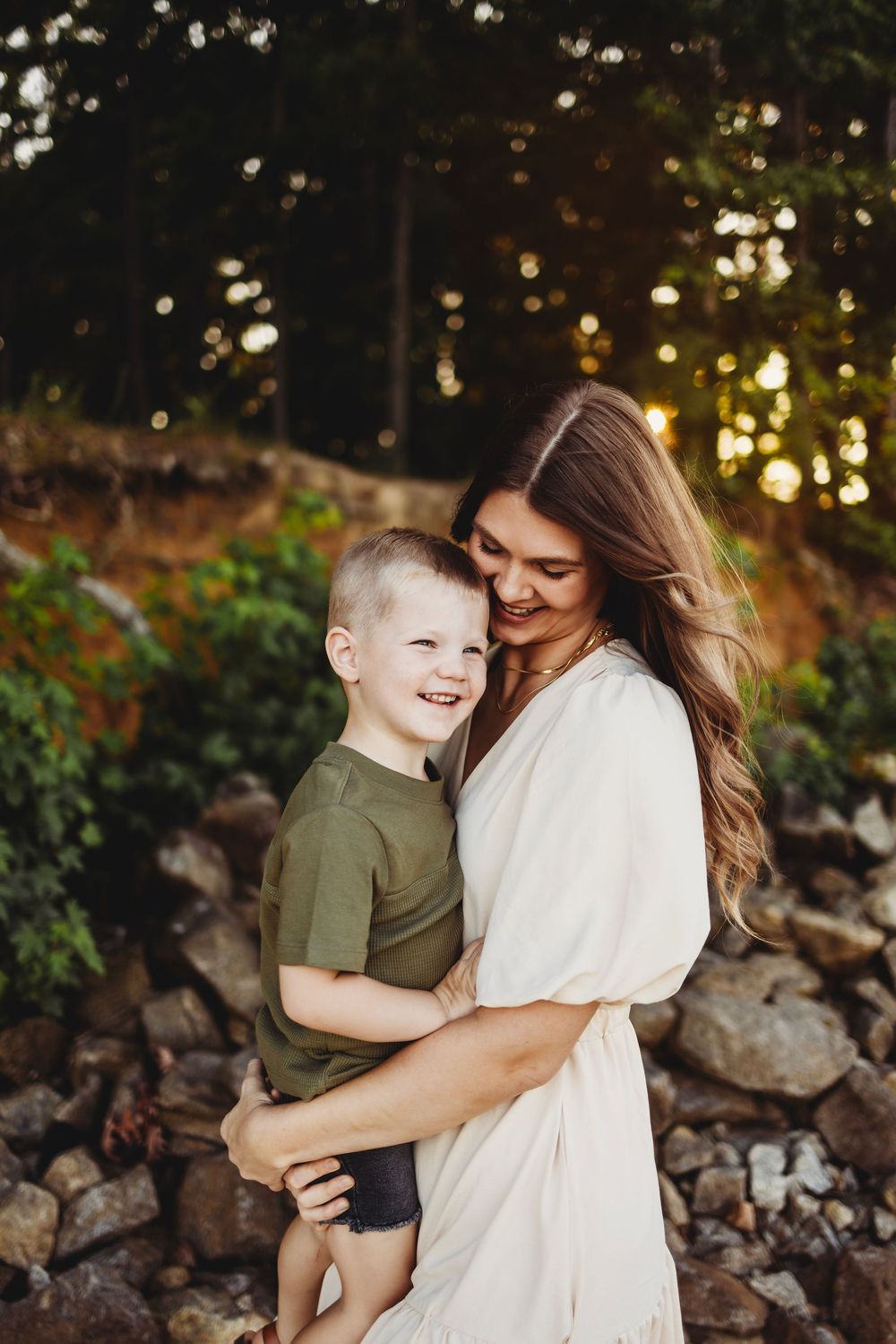 A tender moment of laughter and joy captured outdoors against a sunlit forest backdrop with bokeh lighting effects.