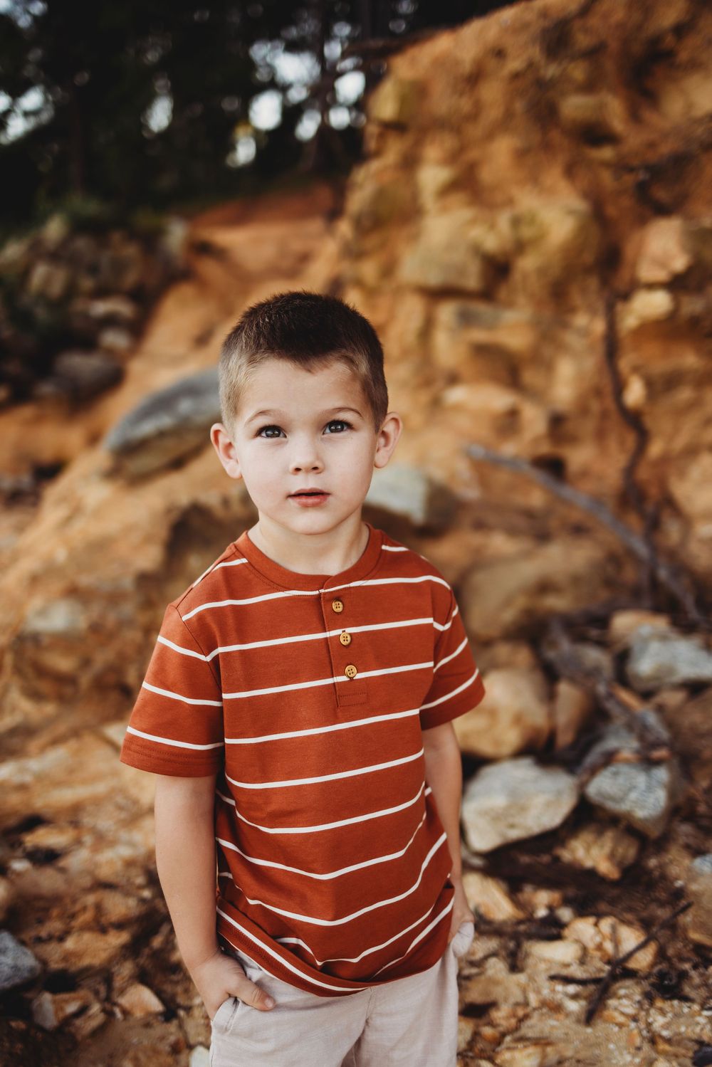 A small child wearing a rust-colored striped polo shirt stands in front of a rocky cliff backdrop on an autumn day.