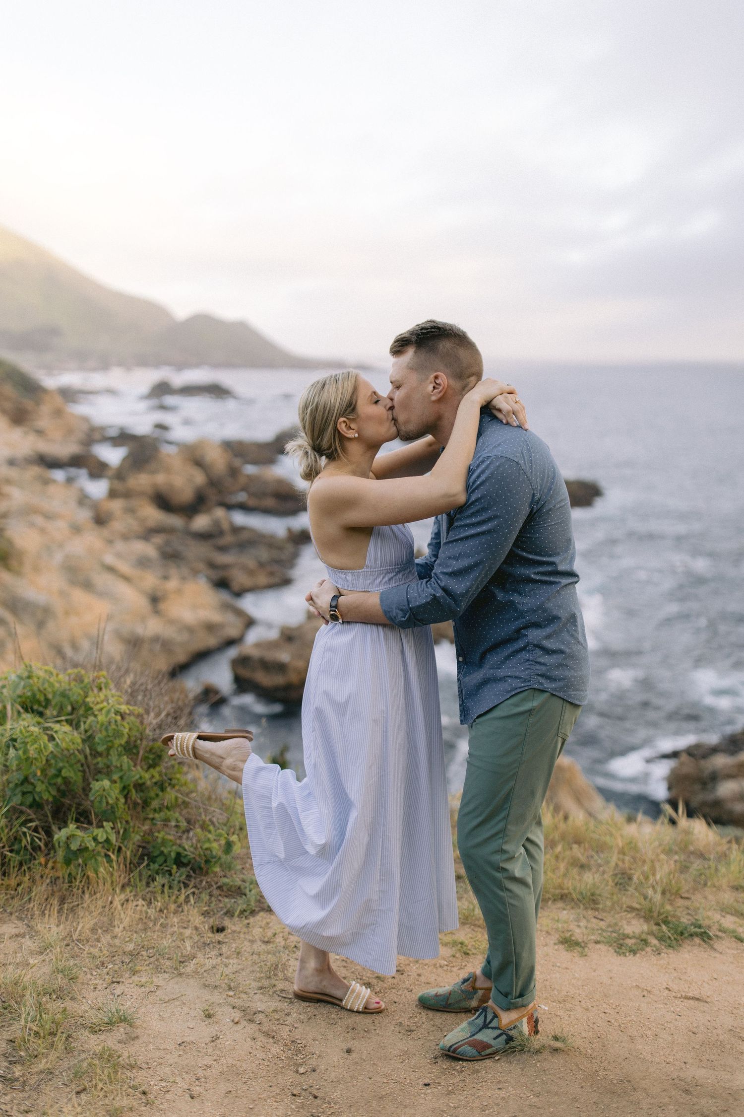 Romantic couple embracing on coastal cliff overlooking rocky shoreline and ocean during golden hour.