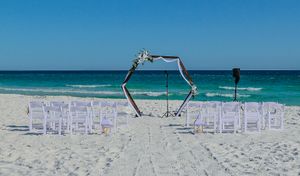 Beach wedding ceremony setup with white chairs and hexagonal arch on pristine sand overlooking turquoise ocean waters.