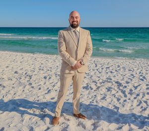 Groom in beige suit standing on white sandy beach with turquoise ocean waves in background.
