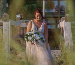Bride in floral lace dress holding bouquet while walking along rustic white fence.