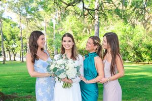 A bridal party in pastel dresses stands on a lush green lawn, with the bride holding a white floral bouquet.