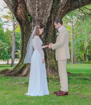 A wedding couple exchanges rings under a large tree in a lush green park setting during their outdoor ceremony.