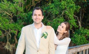 Happy couple poses on wooden bridge during outdoor wedding photo session with green foliage backdrop.